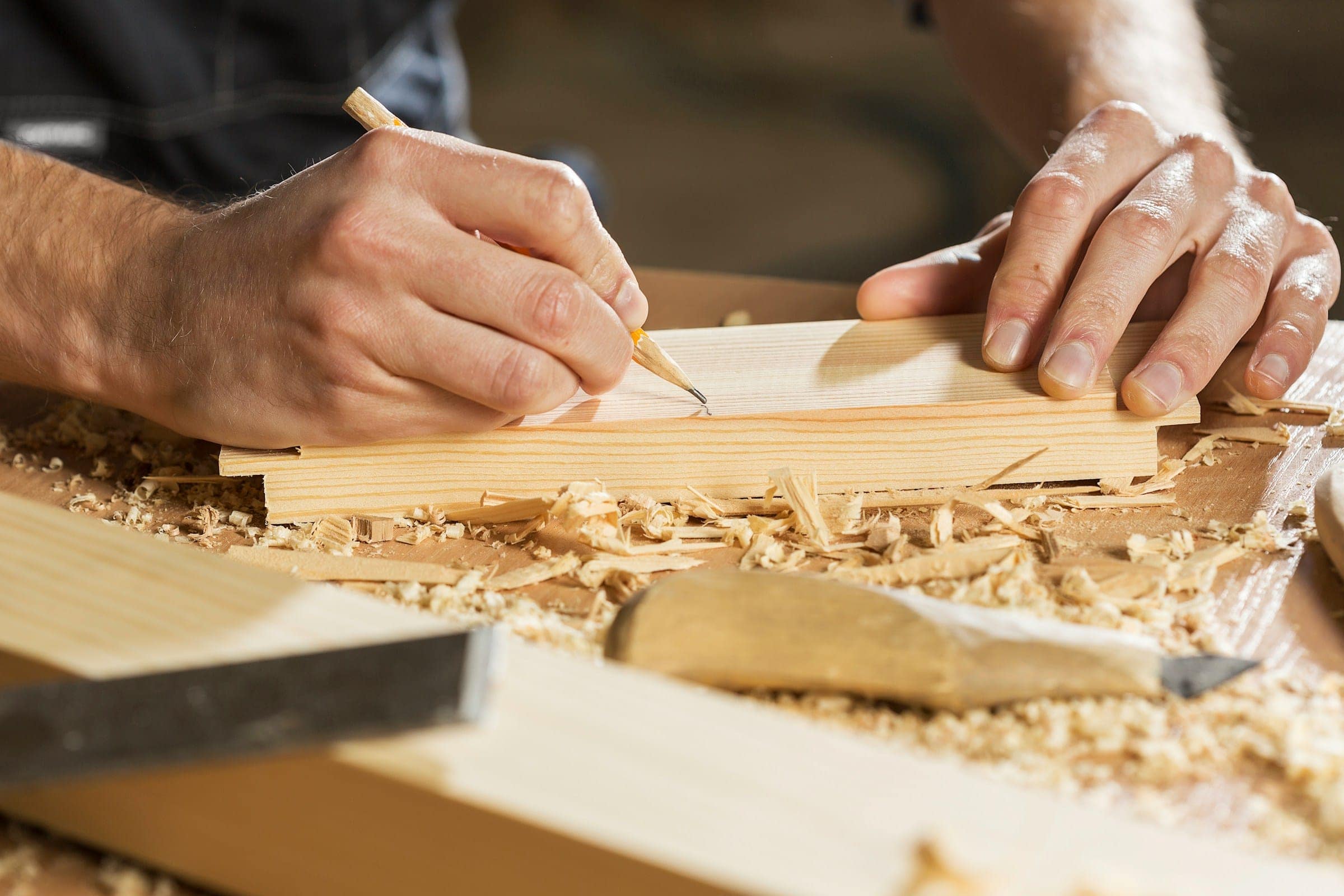 Tischler zeichnet mit Bleistift eine Linie auf ein Holzbrett, daneben liegen Hobelspäne