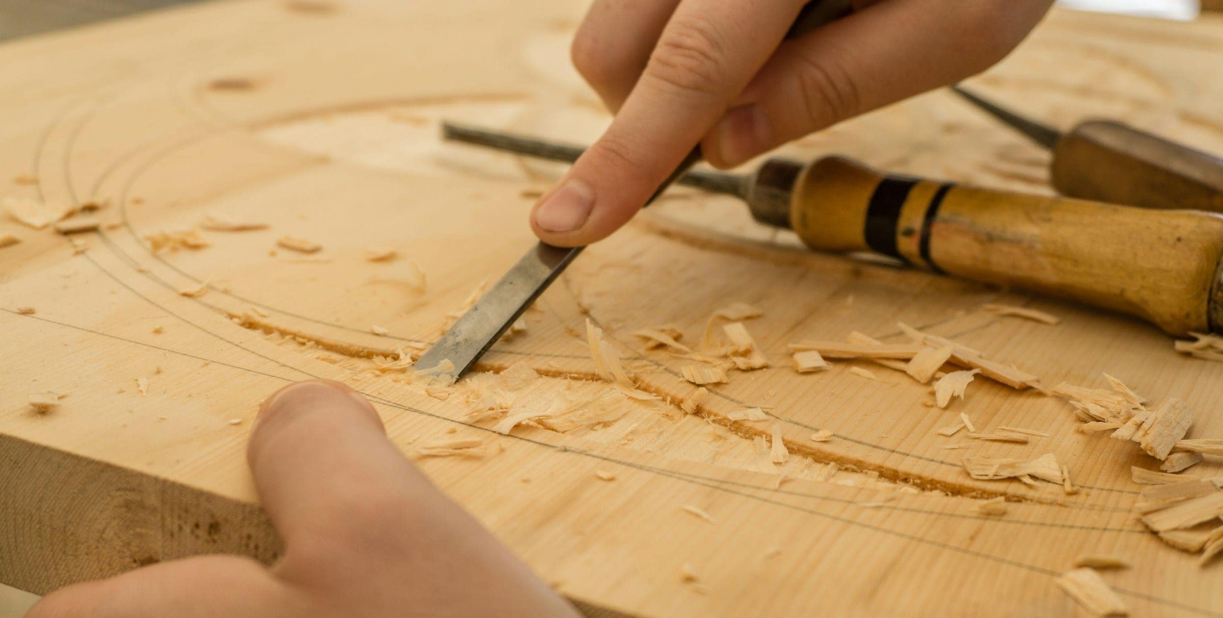 Hand führt einen Stechbeitel durch helles Holz, Späne liegen auf dem Brett